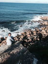 High angle portrait of young man sitting on rock at beach against sky