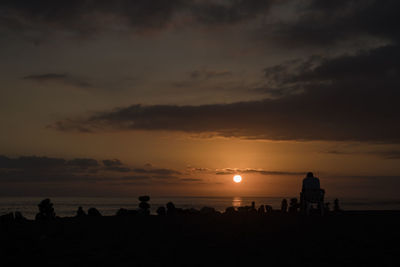 Silhouette man with stacked pebbles at beach against orange sky