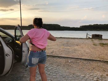 Rear view of woman standing by car against sky during sunset