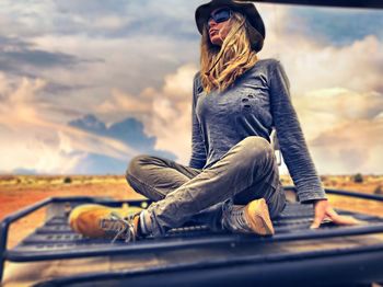 Full length of woman sitting on car roof against cloudy sky