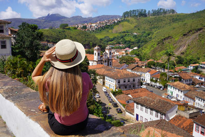 Tourist girl with hat sitting on wall looking at panoramic view of the city of ouro preto, brazil