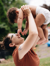 Side view of mother holding baby mid-air standing outdoors