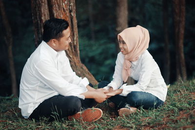 Romantic smiling couple holding hands while sitting on field against trees in forest
