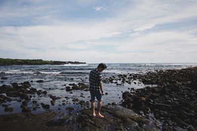 Rear view of man standing on rock at beach against sky