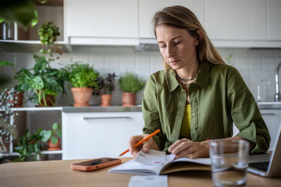 Focused woman attentive budget sitting at table write down expenses in notebook use phone calculator