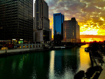 Modern buildings by river against sky during sunset