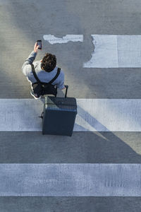 Businessman with baggage crossing the street while looking at cell phone, top view
