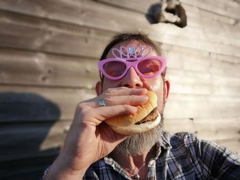 Portrait of man in novelty glasses eating burger against wall
