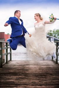 Bride and groom jumping on wooden pier while holding hands