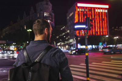 Rear view of man carrying backpack while standing on city street at night