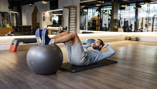 Bearded adult male athlete holding legs on fit ball and doing abdominal crunches on mat during fitness training in gym