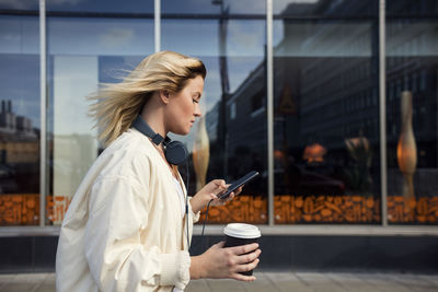 Side view of woman using mobile phone while holding disposable cup against building
