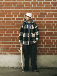 Young man with skateboard posing in front of brick wall