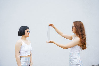 Side view of lesbian woman holding mirror standing with partner against wall