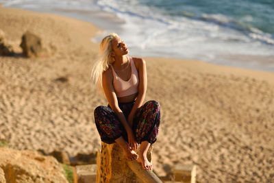 Young woman looking away while sitting on wooden post at beach