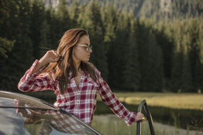 Young woman standing with car against trees