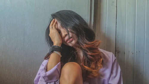 Woman with hand covering face sitting against wall at home