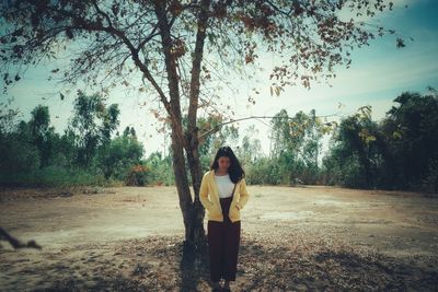 Young woman with hands in pockets standing on field against sky