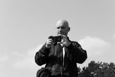 Low angle view of man with shaved head photographing with mobile while standing against sky