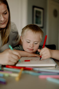 Boy with down syndrome drawing in book by mother at dining table