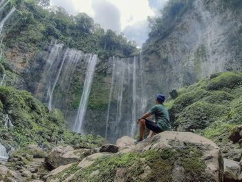 Low angle view of man sitting on rock against waterfall
