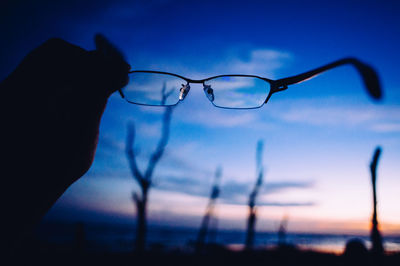 Cropped image of silhouette hand holding eyeglasses against sky during sunset