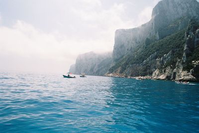 Boats sailing in sea by rocky mountains against sky