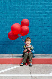Full length of boy with red balloons sitting on sidewalk against blue wall