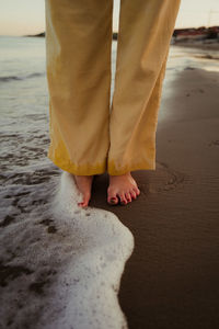 Legs of crop unrecognizable barefoot female standing in foam of wavy ocean in sunset
