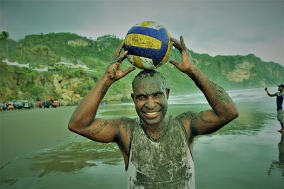 Portrait of messy man holding volleyball while standing on land against mountains and sky