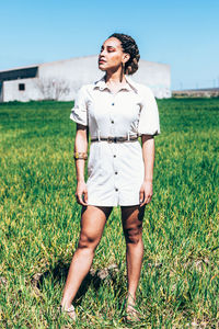 Trendy ethnic female model in casual white dress with traditional african accessories with closed eyes while standing in grassy meadow near farm building