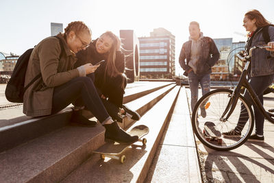 Teenage girls talking on steps while friends standing with bicycle and skateboard in city