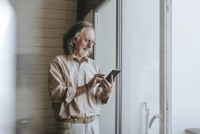 Smiling senior man standing near window using tablet pc at home