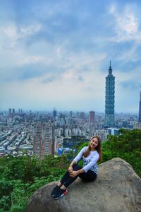 Portrait of young woman sitting on modern cityscape against sky