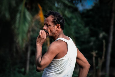 Side view of man smoking cigarette while standing against trees