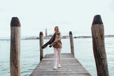 Portrait of young woman walking on pier over river against historic buildings