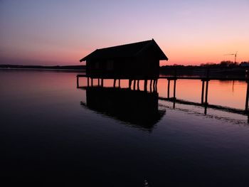 Silhouette wooden house over lake against sky during sunset
