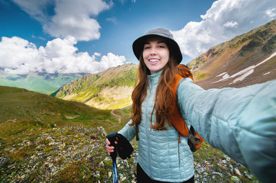 Smiling young woman takes a selfie on a mountain peak, with a backpack on a sunny day. tourist