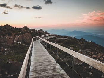 Footbridge leading towards mountains against sky during sunset