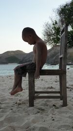 Side view of shirtless boy sitting on chair at beach