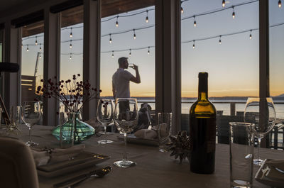 Man seen through window drinking wine while standing against sky during sunset