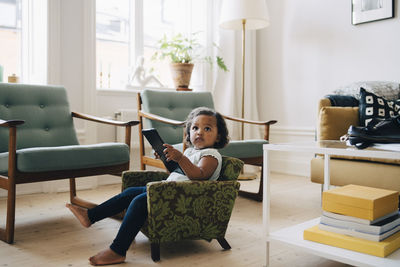 Full length of girl holding digital tablet while sitting on small armchair at home