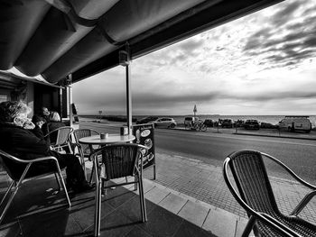 Empty chairs and table by window against sky