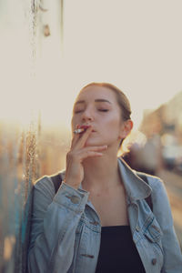 Young woman smoking while standing by wall in city against sky