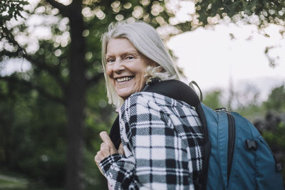 Portrait of happy senior woman looking over shoulder carrying backpack while hiking