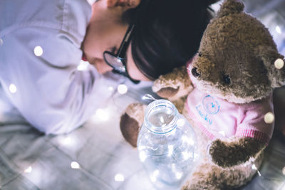 High angle view of young woman sleeping by teddy bear and string lights at home