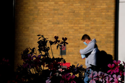 Man  with face mask walking brick wall  by flowering plants against wall