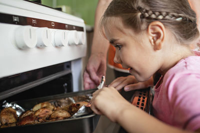 Excited small girl and caring grandmother taking pan with baked roast chicken and potato out of oven