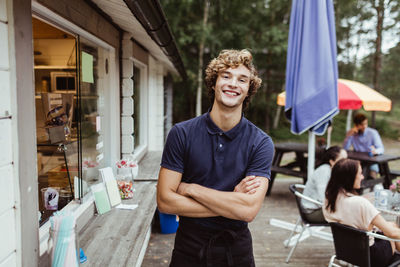 Portrait of young owner with arms crossed standing by concession stand while customer sitting in background