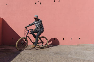 Confident man riding bicycle on sidewalk against peach wall during sunny day